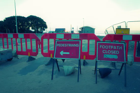 A photograph of Exmouth seafront with barriers across the footpath. Signs direct pedestrians to cross the road and say "footpath closed".