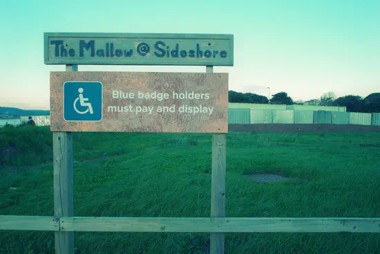 A photograph of The Mallow at Sideshore on Exmouth seafront. A sign reads "The Mallow @ Sideshore" and "Blue badge holders must pay and display" with The Mallow (a field) behind it. The furthest area of The Mallow is fenced off for the sea wall replacement works.