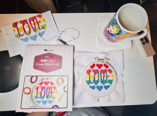 A photograph of a desk set-up for cross stitching. At the top-left is a pattern of coloured hearts with “love” printed in the centre. At the bottom-left is the Hobbycraft box that the kit arrived in. At the top-right is a coffee-stained Wonder Woman mug. At the bottom-right is the complete cross stitch.