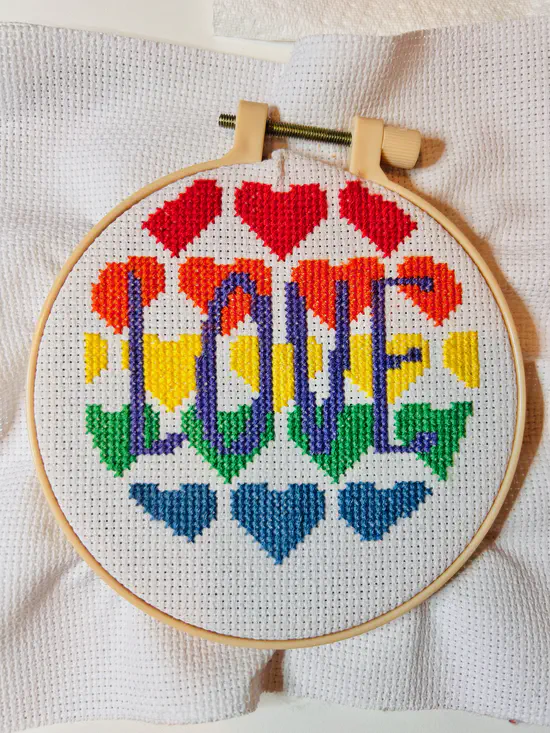 A photograph of a completed cross stitch. From top to bottom are rows of red, orange, yellow, green and blue hearts. The word “love” is printed in purple in the centre.