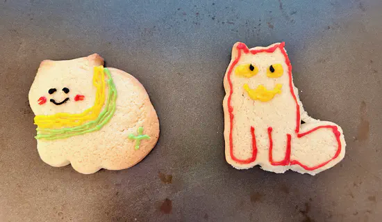 A photograph of two cat-shaped biscuits. The cat on the left is smiling and wearing a green-yellow scarf. The cat on the right has a red outline with yellow eyes and a demonic yellow smile.