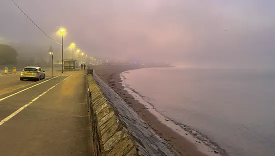 A photograph of Exmouth Beach early in the morning. The street lights are on and illuminating the fog that obscures the distance.