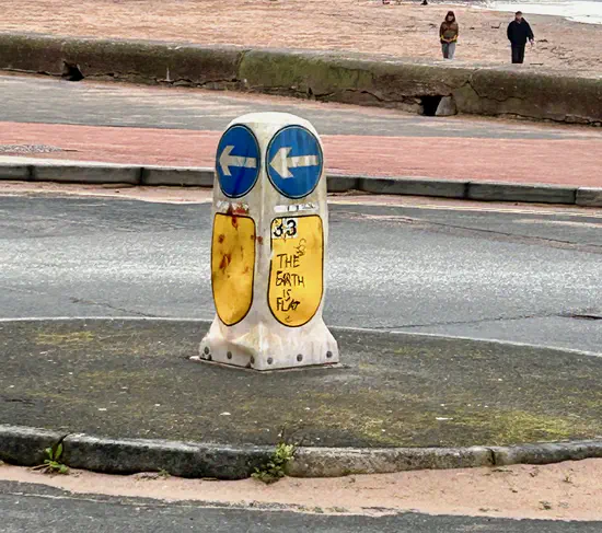 A photograph of a traffic bollard on Exmouth seafront. Graffiti on it reads 'The Earth is Flat'.